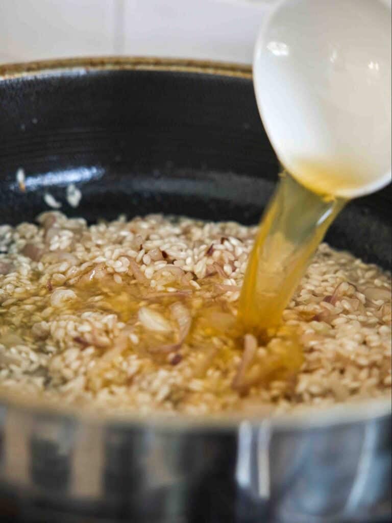 A close-up of a ladle pouring broth into a pan of rice, likely in the process of making risotto, with visible grains and pieces of onion or shallot.